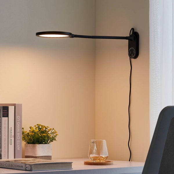 Moderne schwarze LED-Wandleuchte über einem Schreibtisch mit Büchern, kleiner Pflanze und einem Glas in einem hellen Arbeitsbereich.