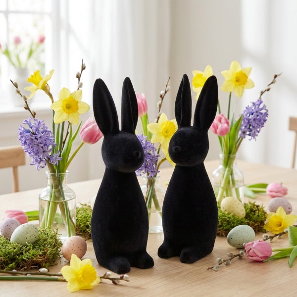 Zwei Osterhasen aus beflocktem Material als Tischdekoration umgeben von Frühlingsblumen und Ostereiern auf einem Holztisch.