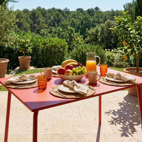 Moderner roter Gartentisch aus Metall, gedeckt auf einer sonnigen Terrasse mit Blick auf den Wald.