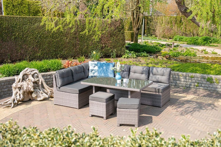 Garten-Lounge-Set aus grauem Polyrattan mit Ecksofa, zwei Hockern und Tisch mit Glasplatte auf einer gepflasterten Terrasse im Garten.