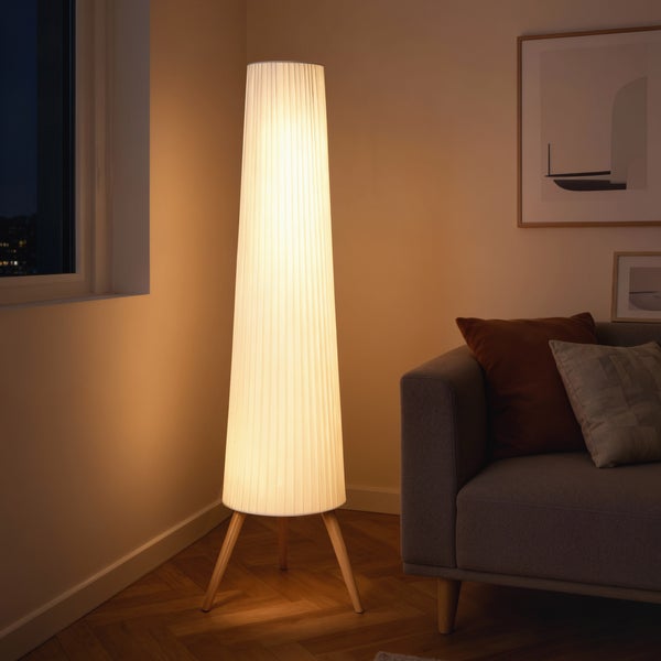 Stehleuchte mit konischem, plissiertem Textilschirm und Holz-Dreibeinstativ in einem warm beleuchteten Wohnzimmer neben einem Sofa.