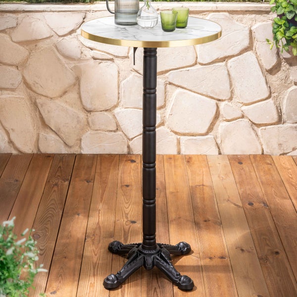 Bistrotisch mit runder Platte in Marmor-Optik, Goldrand und schwarzem Metallfuß auf einer Holzterrasse vor einer Steinwand.