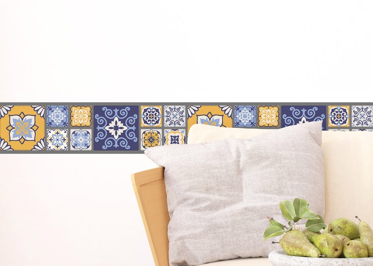Selbstklebende Bordüre in Fliesenoptik mit mediterranen Mustern an einer Wand in einem hellen Wohnraum mit Sessel und Kissen.