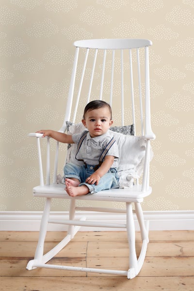 Ein Baby sitzt in einem weißen Schaukelstuhl vor einer Wand mit Wolkenmuster.