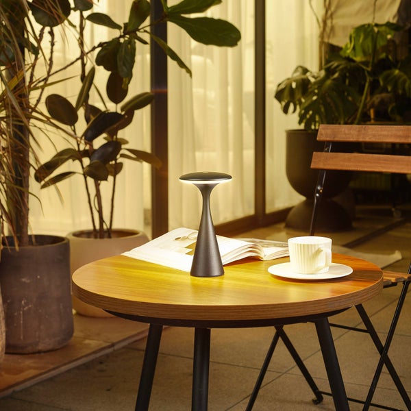 Moderne schwarze Tischleuchte auf einem runden Holztisch mit einem Buch und einer Tasse, umgeben von Zimmerpflanzen in einem hellen Raum.