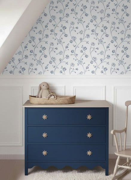 Vliestapete mit blauem Blumenmuster in einem Kinderzimmer über weißer Wandpaneele und einer dunkelblauen Kommode.