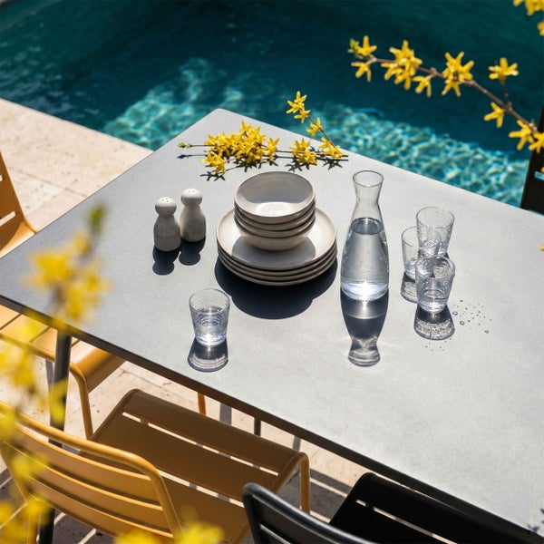 Moderner grauer Gartentisch mit gelben und schwarzen Metallstühlen auf einer sonnigen Terrasse am Pool, gedeckt mit Geschirr und Gläsern.