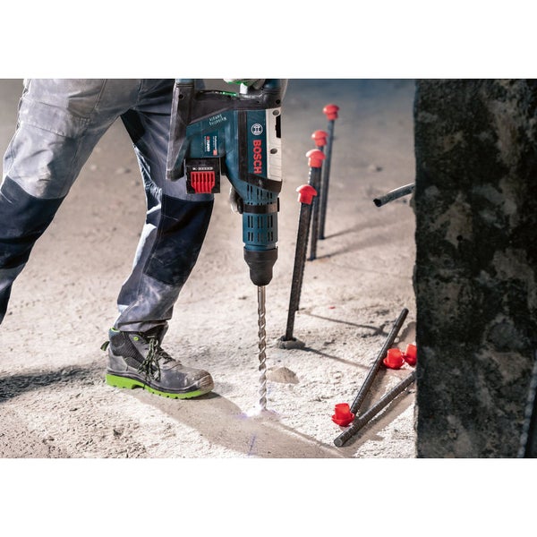 Ein Mann bohrt mit einem Bohrhammer Löcher in einen Betonboden. Im Hintergrund sind Armierungseisen zu sehen. Bosch Logo.