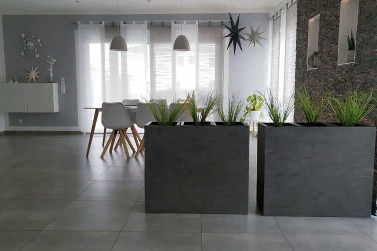 Zwei große rechteckige anthrazitfarbene Pflanzkübel mit Ziergras in einem modernen hellen Wohnbereich mit grauen Bodenfliesen und Steinwand.