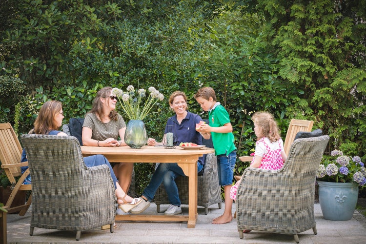Familie sitzt an einem runden Holztisch im Garten auf geflochtenen Sesseln und genießt einen Nachmittag mit Obst.