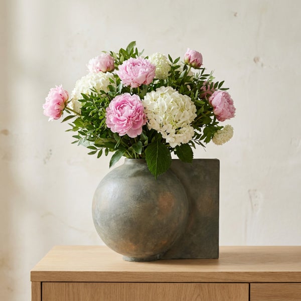 Blumenstrauß aus Pfingstrosen und Hortensien in einer modernen, kugelförmigen Steinvase auf einem Holzmöbelstück.