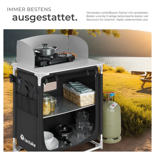 Campingküchenschrank mit Windschutz, Kochfeld und Stauraum vor einer Naturkulisse.