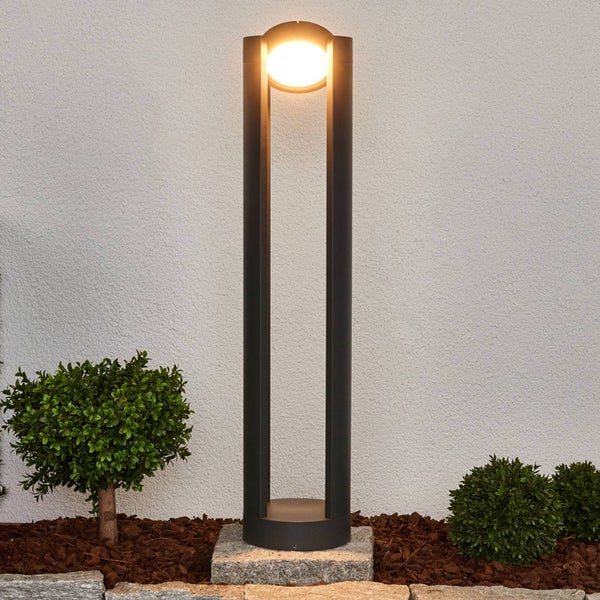 Moderne LED-Pollerleuchte aus Metall in Anthrazit mit warmweißem Licht in einem organisierten Gartenbeet vor einer weißen Wand.