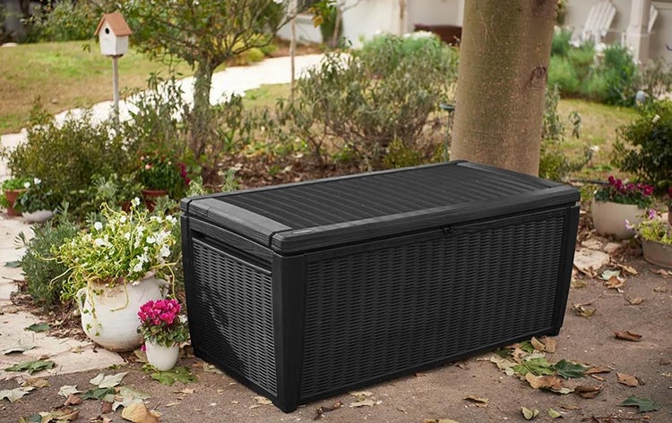 Große schwarze Garten-Aufbewahrungsbox mit Flechtstruktur in einem hellen Garten mit Blumentöpfen und Bäumen.