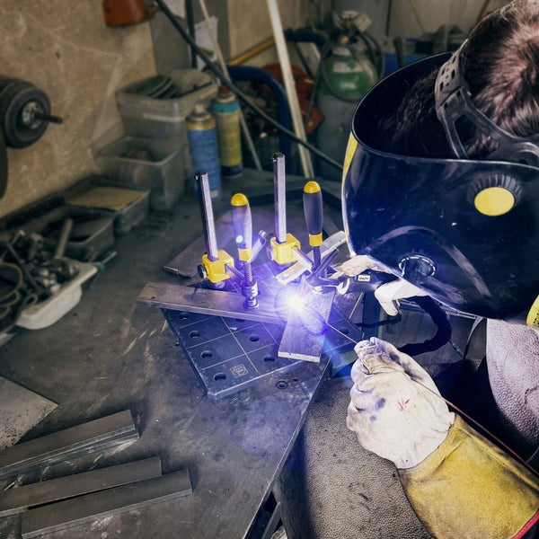Person beim Schweißen von Metallteilen mit Schutzmaske und Handschuhen auf einem Schweißtisch mit Schraubzwingen.