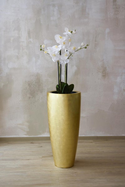 Weiße künstliche Orchidee in einer hohen goldfarbenen Bodenvase vor einer strukturierten Wand.