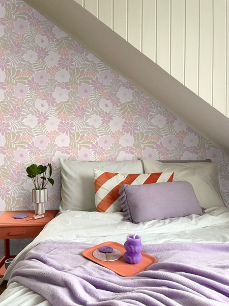 Hell eingerichtetes Schlafzimmer mit floral gemusterter Tapete an einer Dachschräge, einem Bett mit Kissen und einem Nachttisch mit Zimmerpflanze.