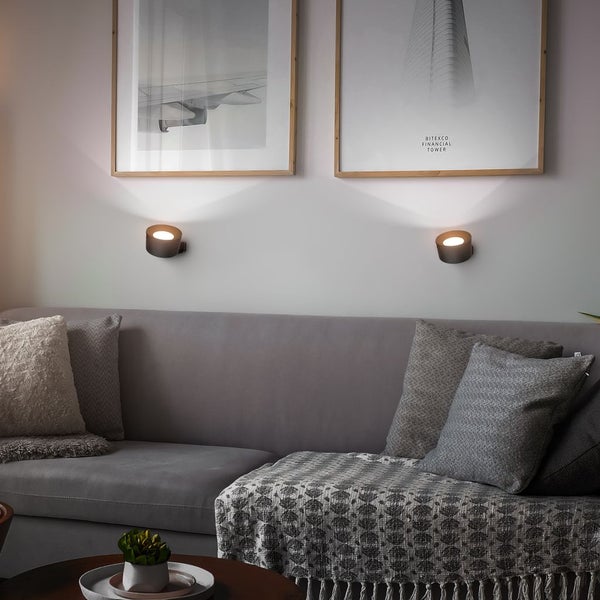 Zwei moderne, runde Wandleuchten in Schwarz über einem grauen Sofa im Wohnzimmer, indirekte Beleuchtung unter gerahmten Bildern.