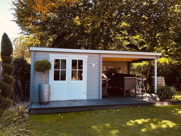 Gartenhaus aus Holz mit Flachdach, Doppeltür mit Glaseinsatz und integriertem Terrassenanbau mit Außenküche und Grill auf einer Holzterrasse.