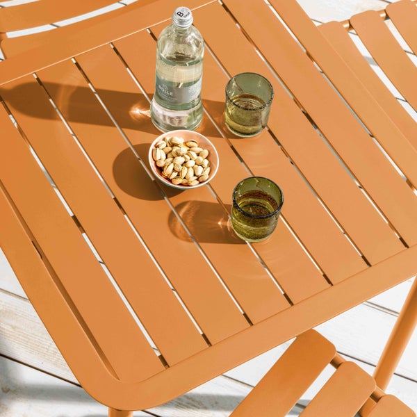 Gartentisch aus Metall mit Lattenstruktur, darauf eine Wasserflasche, Gläser und eine Schale Pistazien auf einer Holzterrasse.