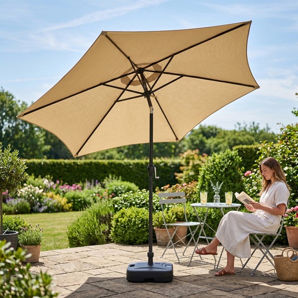Beiger quadratischer Sonnenschirm mit schwarzem Mast und Standfuß auf einer Terrasse im Garten mit Sitzgruppe und einer lesenden Frau.
