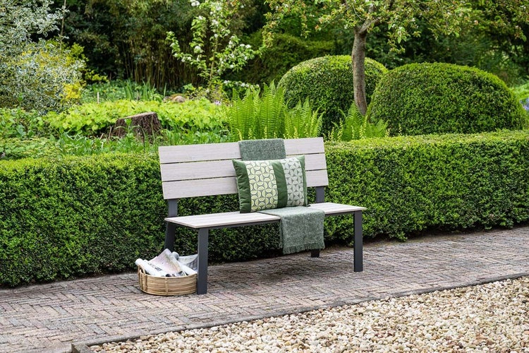 Moderne Gartenbank aus hellen Holzlamellen und dunklem Metall in einer hellen, gepflegten Gartenanlage mit Hecken und gepflastertem Weg.