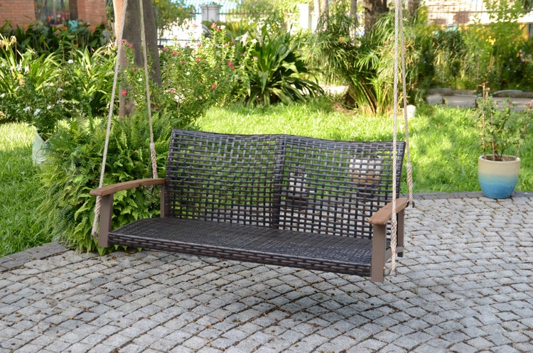 Garten-Schaukelbank aus dunklem Rattangeflecht mit Armlehnen aus Holz, hängend an Seilen in einem Garten über einer gepflasterten Fläche.