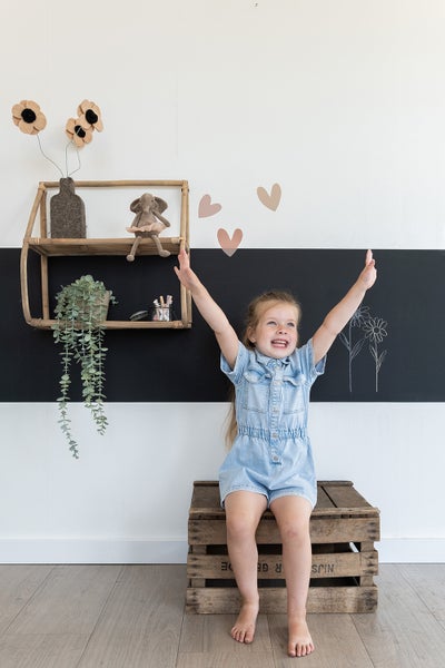 Fröhliches Mädchen sitzt auf einer Holzkiste in einem verspielten Kinderzimmer mit Regalen und Wanddekoration