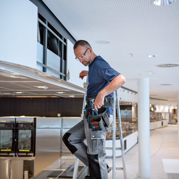 Ein Handwerker mit Werkzeuggürtel steht auf einer Leiter in einem gewerblichen Küchenbereich und führt Installationsarbeiten durch.