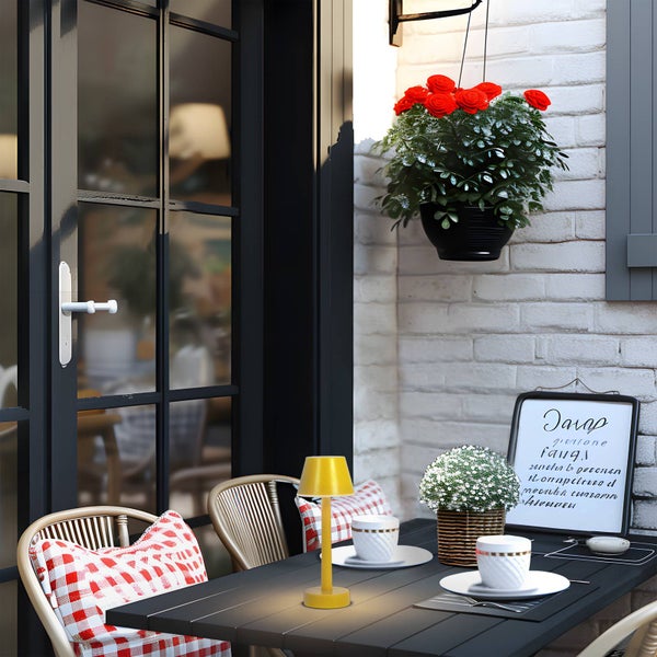Tragbare gelbe Tischleuchte auf einem dunklen Gartentisch mit Stühlen, Kissen und einer hängenden Blumenampel auf einer gemütlichen Terrasse.