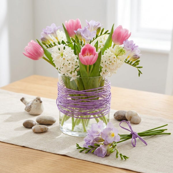 Frühlingsstrauß mit Tulpen, Hyazinthen und Freesien in einer Glasvase, dekoriert auf einem Holztisch mit Dekosteinen und einer kleinen Figur.