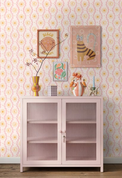 Wohnraumszene mit gemusterter Tapete mit Sonnen- und Mondmotiven zwischen Wellenlinien. Ein rosa Glasschrank mit geriffeltem Glas steht vor der Wand.