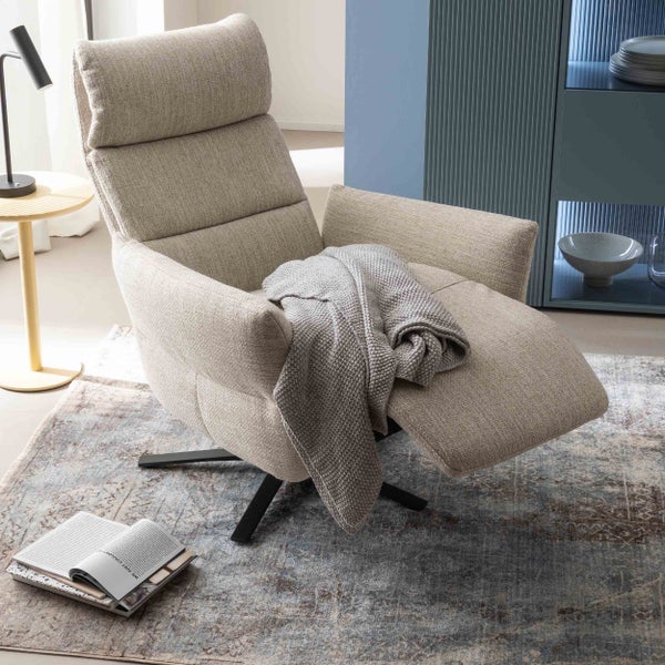 Moderner Relaxsessel aus beigem Strukturstoff mit schwarzem Metalldrehfuß, grauer Strickdecke und Teppich in einem hellen Wohnzimmer.