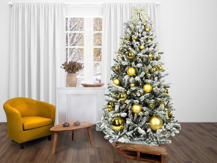 Geschmückter künstlicher Weihnachtsbaum mit Schneeoptik und goldenen Weihnachtskugeln in einem Wohnzimmer mit Sessel und kleinem Holztisch.