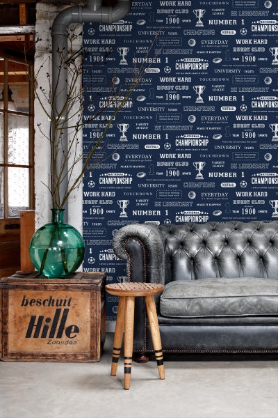 Innenraum mit Sofa, Hocker, Holzkiste und Vase vor einer mit sportlichen Motiven bedruckten Tapete