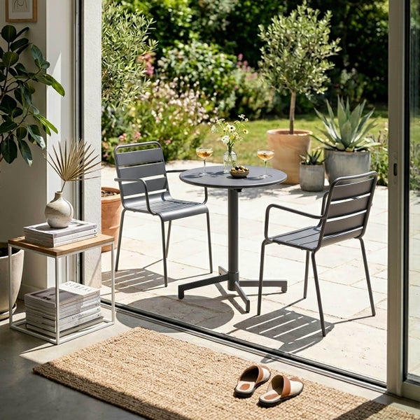Gartenset bestehend aus einem runden grauen Metalltisch und zwei grauen Metallstühlen auf einer sonnigen Terrasse mit Pflanzen im Hintergrund.