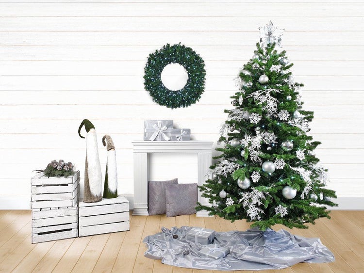 Geschmückter Weihnachtsbaum mit silberner Dekoration, beleuchtetem Adventskranz, zwei Zwergfiguren und Geschenkpaketen vor weißer Wand.