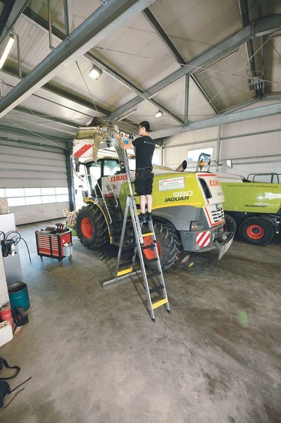 Ein Servicetechniker reinigt den Auswurfkrümmer eines Claas Jaguar Feldhäckslers auf einer hohen Arbeitsplattform in einer landwirtschaftlichen Halle.