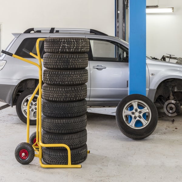 Gelbe Reifenkarre mit acht Reifen in einer hellen Autowerkstatt mit einem silbernen Auto auf einer Hebebühne.