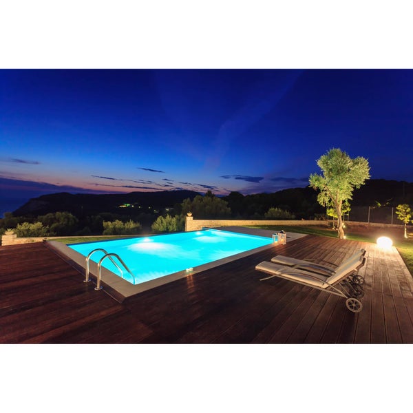 Beleuchteter rechteckiger Pool bei Nacht mit dunkler Holzterrasse, zwei Sonnenliegen und stimmungsvoller Außenbeleuchtung.