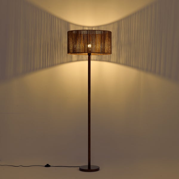 Eingeschaltete Stehlampe mit zylindrischem Schirm aus feinen Fäden, die ein warmes Licht und dekorative Streifenschatten an die Wand wirft.