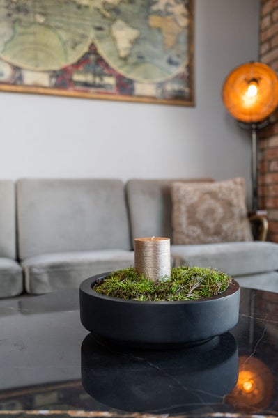 Dekoratives Gesteck aus Schale, Moos und metallischer Stumpenkerze auf einem Tisch in einem Wohnzimmer mit Sofa und Stehlampe.