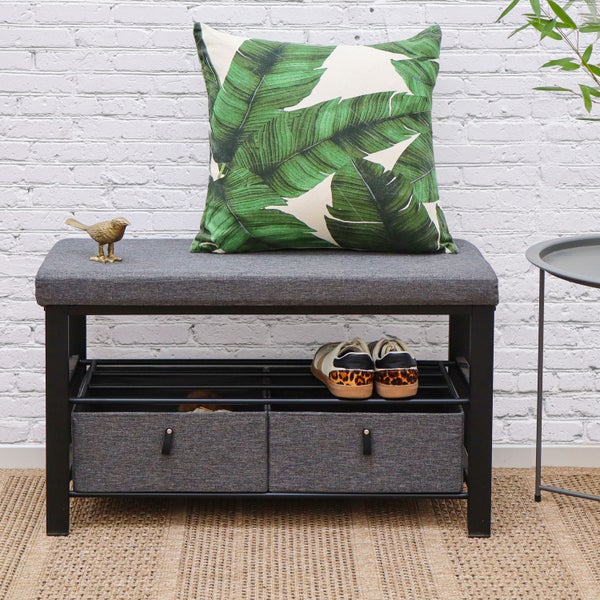 Gepolsterte Schuhbank mit Metallgestell, Schuhablage und zwei Stoffschubladen vor einer weißen Ziegelwand.