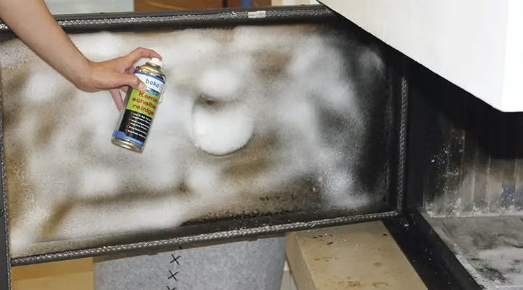 Eine Hand sprüht beko Kaminglasscheibenreiniger auf die verrußte Glasscheibe einer Kaminofentür.
