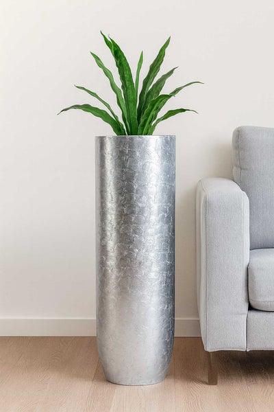 Hohes zylindrisches Pflanzgefäß in Silber mit strukturierter Oberfläche und grüner Pflanze neben einem Sofa auf hellem Holzboden.