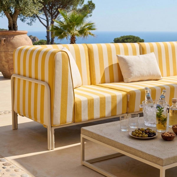 Gestreiftes Gartensofa mit Metallgestell und Steincouchtisch auf einer sonnigen Terrasse mit Meerblick, Olivenbaum und Erfrischungen.