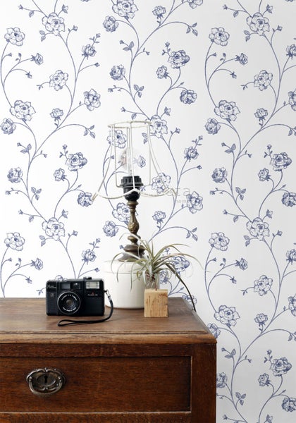 Tapete mit blauem Blumenmuster auf weißem Hintergrund, dargestellt in hellem Raum mit Vintage-Kamera und Lampe auf einer Holzkommode.