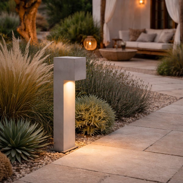 Moderne Sockelleuchte aus Beton in einem Garten mit Kiesweg und Bepflanzung bei Abendstimmung für eine stimmungsvolle Außenbeleuchtung.