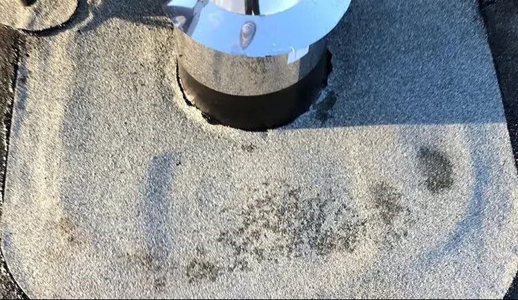 Nahaufnahme einer Dachdurchführung mit Metalleinfassung auf einer mit Bitumen abgedichteten Dachfläche.