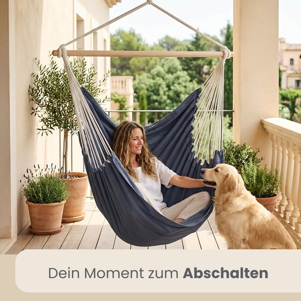 Frau sitzt entspannt in einem blauen Hängesessel auf einem hellen Balkon mit Holzfußboden, einem Hund und verschiedenen Topfpflanzen.